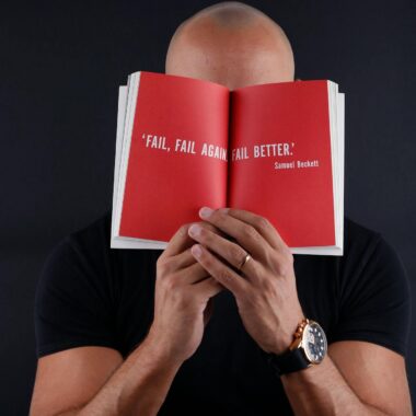 Bald man with wristwatch holding open book displaying inspirational quote by Samuel Beckett.