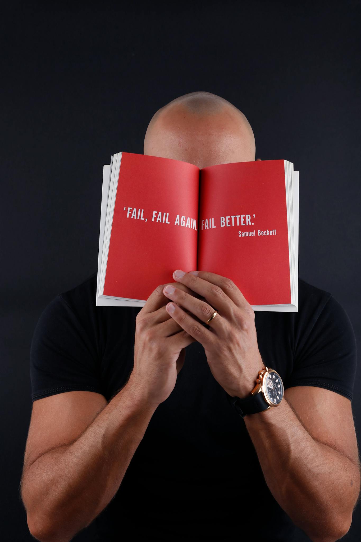 Bald man with wristwatch holding open book displaying inspirational quote by Samuel Beckett.