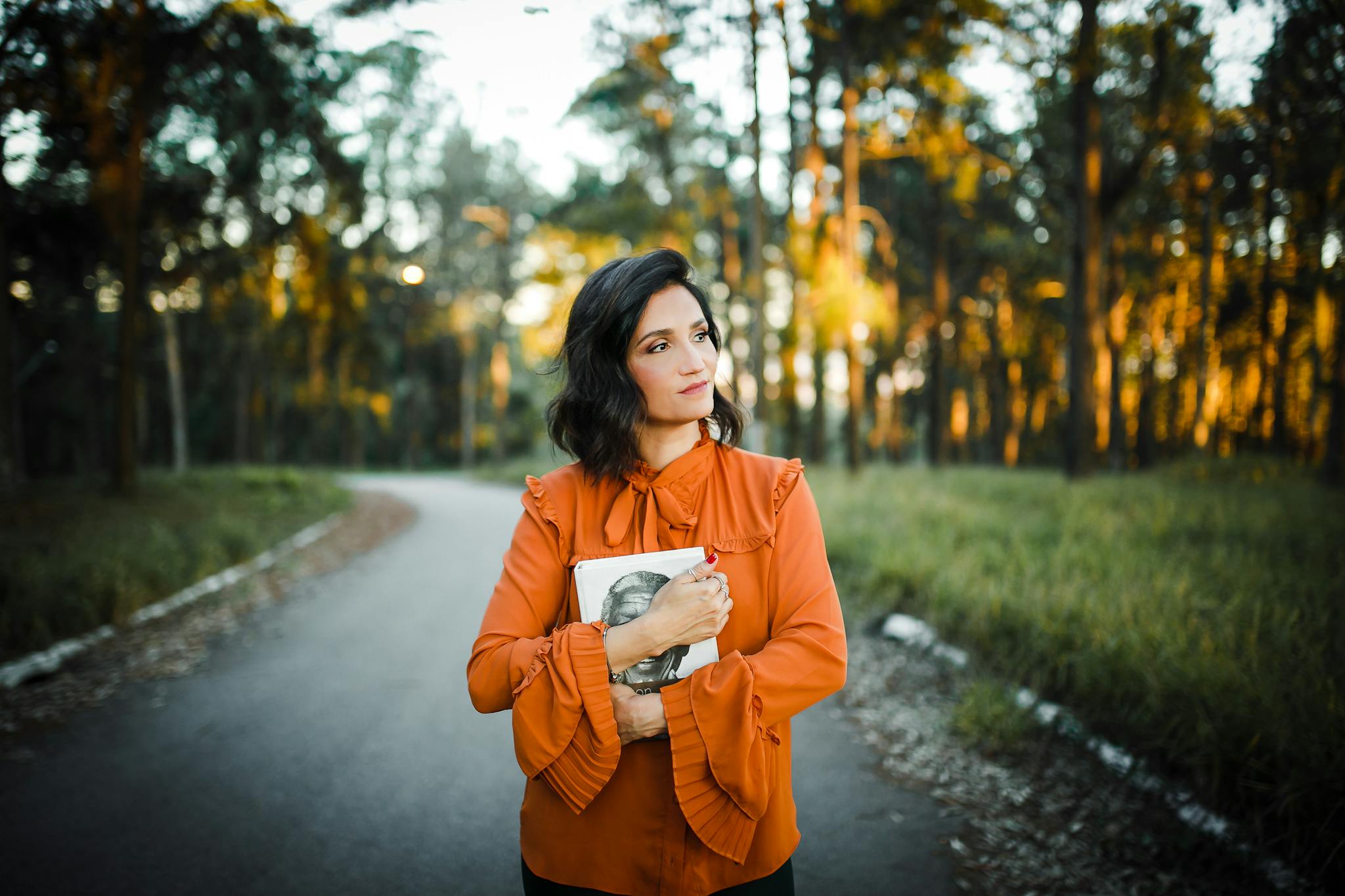 Elegant woman in orange blouse holding book on forest road at sunset.